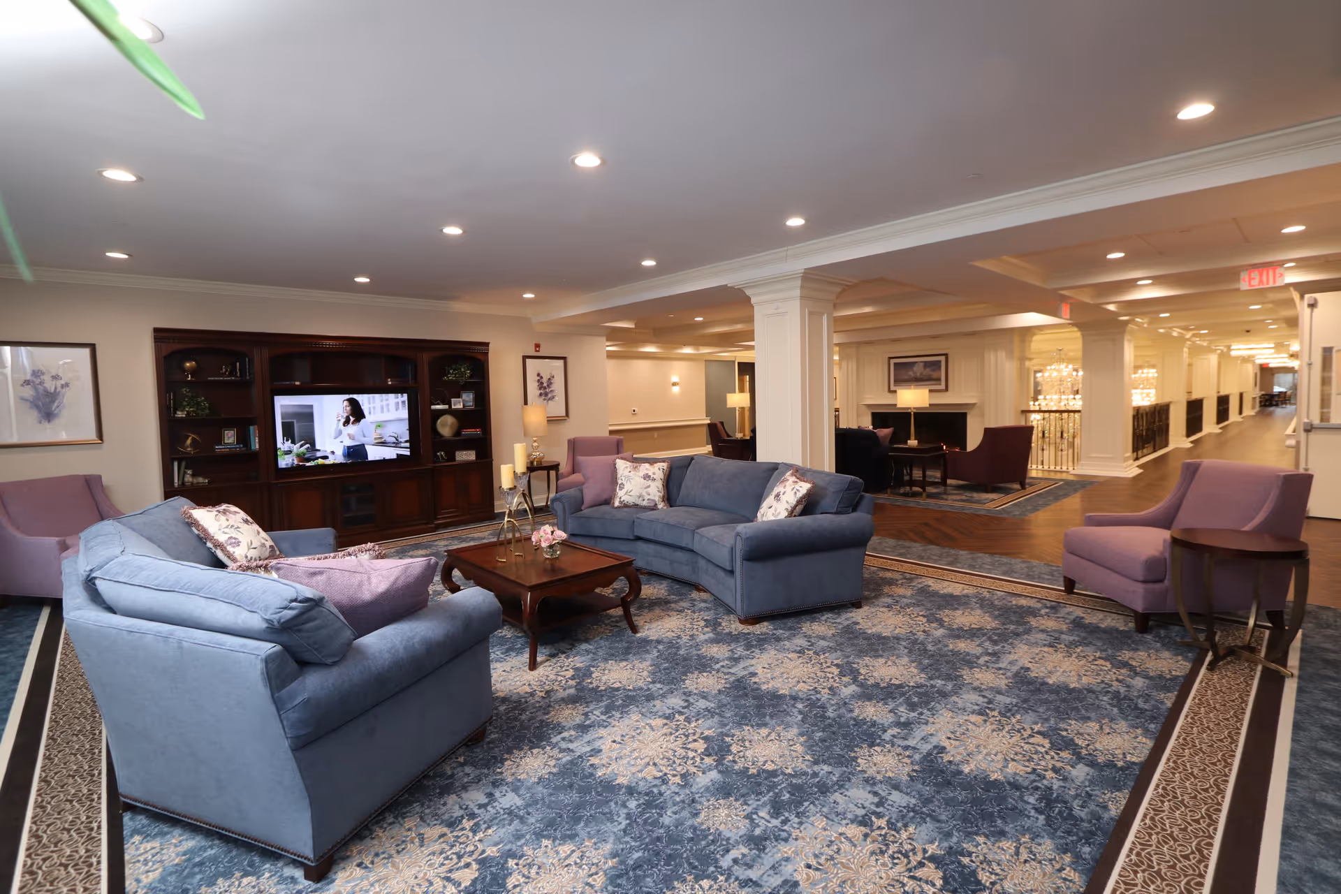 A spacious and well-lit living room area with blue and purple upholstered sofas and armchairs arranged around a wooden coffee table on a patterned blue carpet. A large wooden entertainment center with a television is visible against the wall, and framed artwork and lamps decorate the space. The room opens into a hallway with additional seating areas and elegant lighting fixtures.
