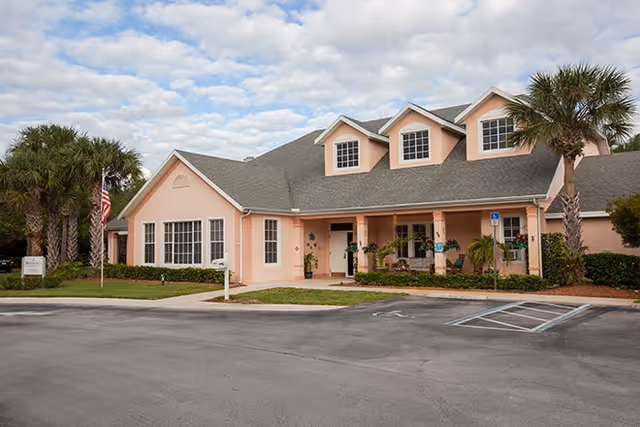 Pink senior living facility building with a covered entrance, palm trees, and a parking lot.