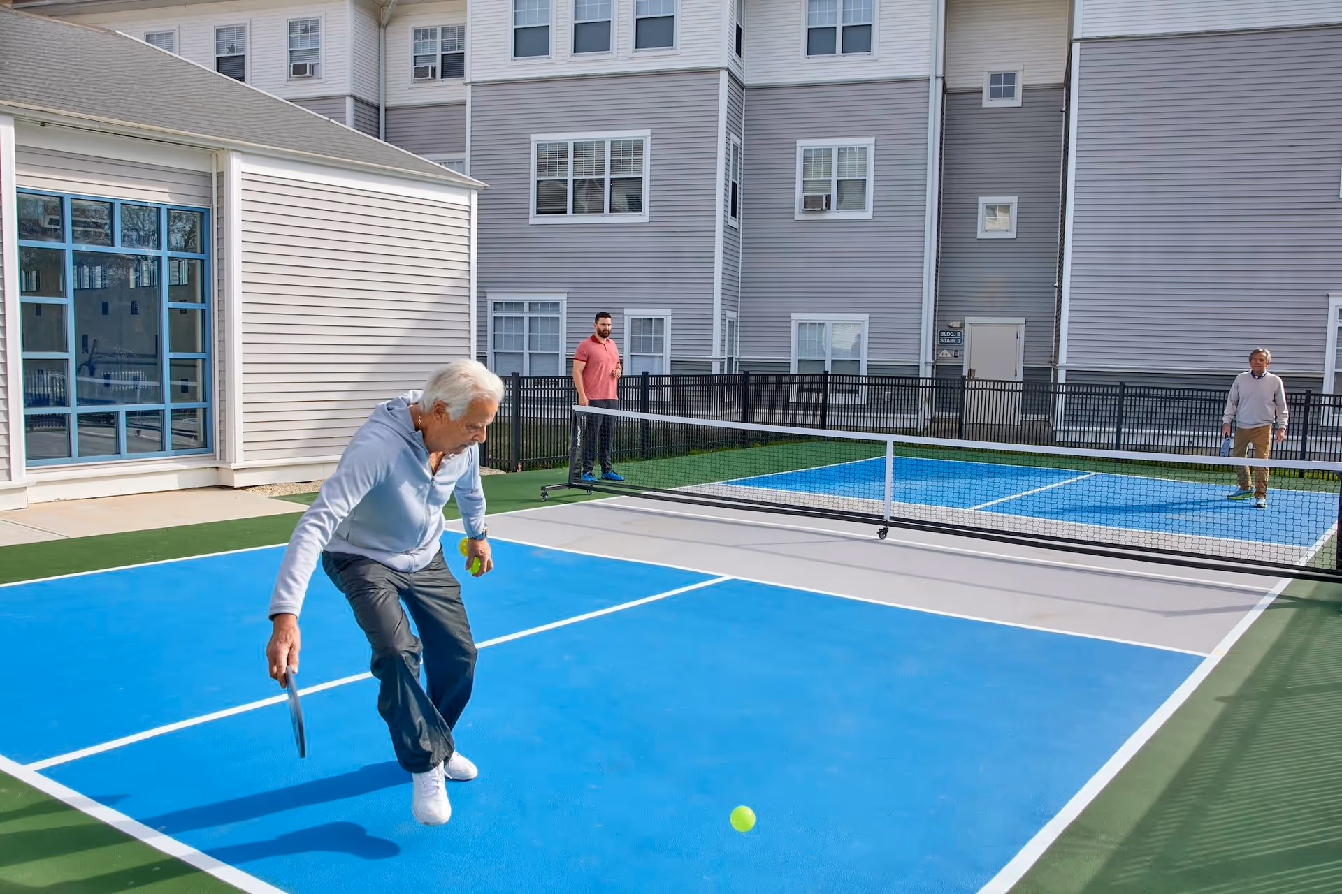 Three people play pickleball on a blue outdoor court next to a multi-story residential building.
