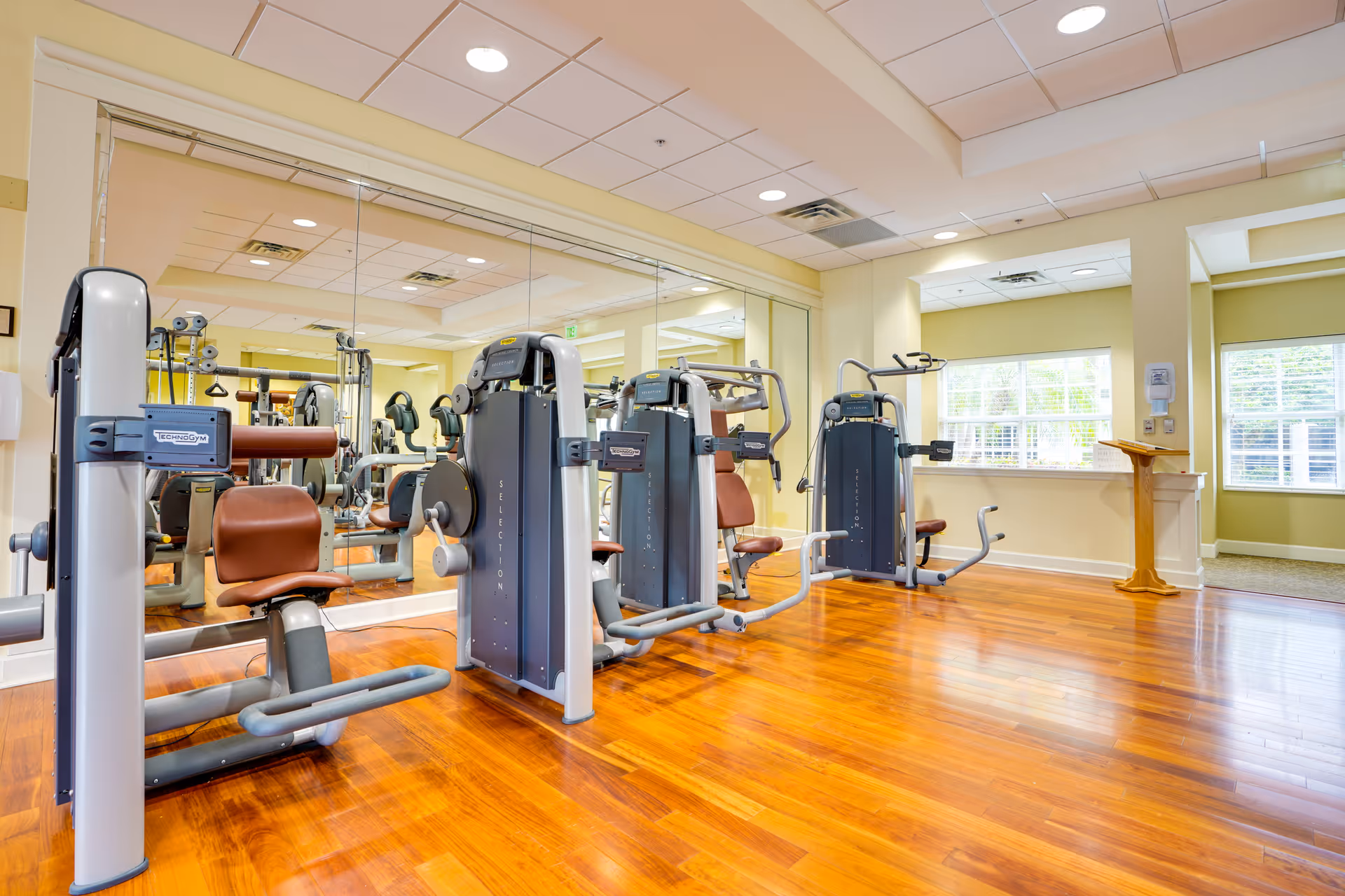 Bright fitness room with wooden floors and several exercise machines positioned in front of a large wall mirror. The room has light yellow walls, a white ceiling with recessed lighting, and windows letting in natural light.