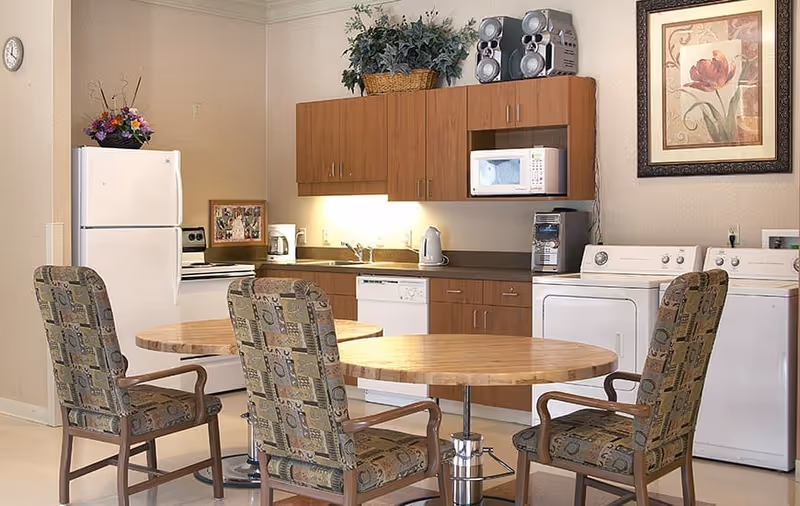 A kitchen and laundry area with wooden cabinets, a white refrigerator, stove, microwave, dishwasher, washing machine, and dryer. There is a round wooden table with four patterned upholstered chairs around it. A framed floral picture hangs on the wall, and a basket with green plants is placed on top of the cabinets.