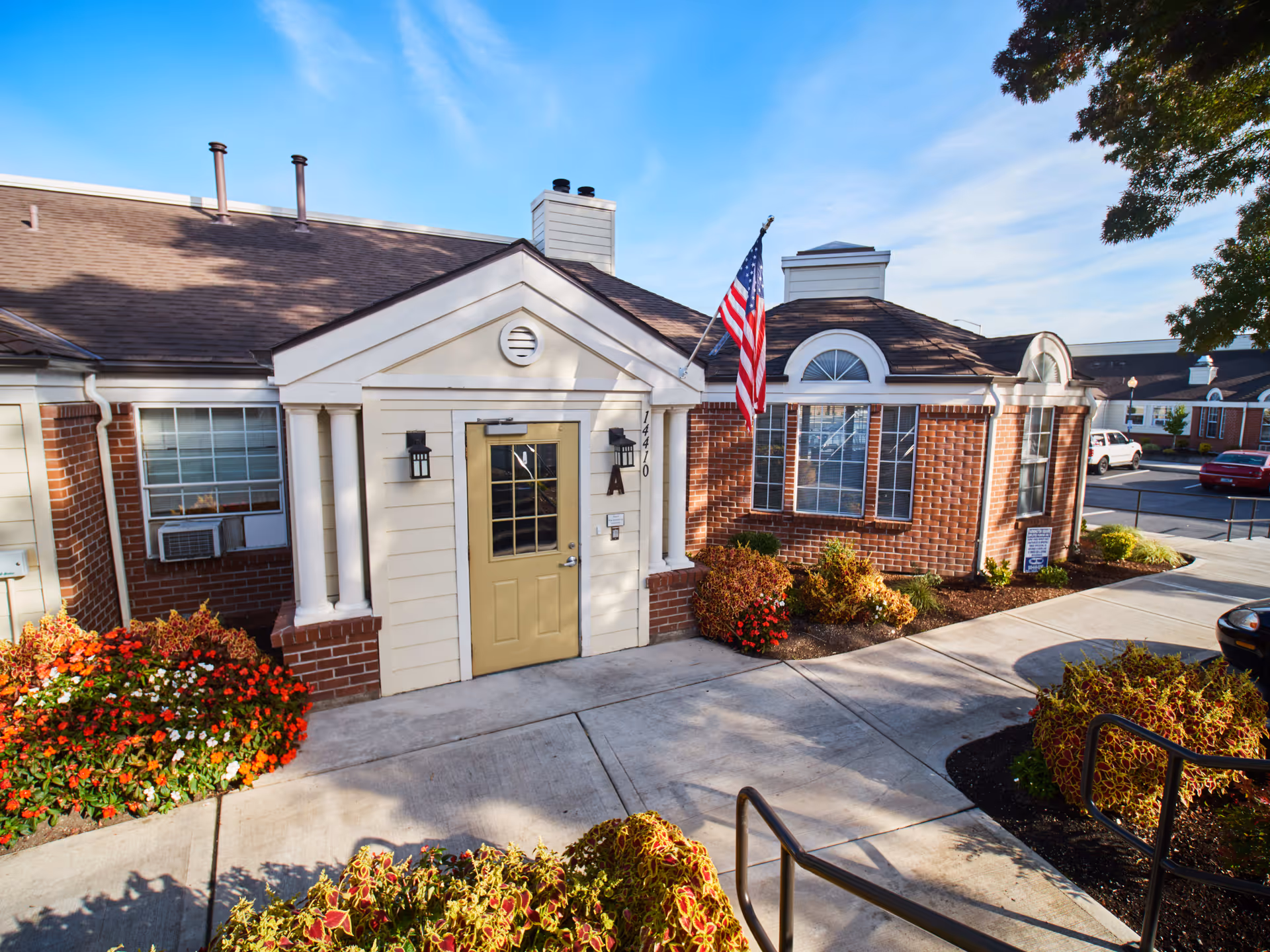 Exterior view of a brick and siding building with a beige door labeled 'A' and the number 14410. There is an American flag mounted near the door, flower beds with colorful flowers and shrubs, a concrete walkway, and a clear blue sky.