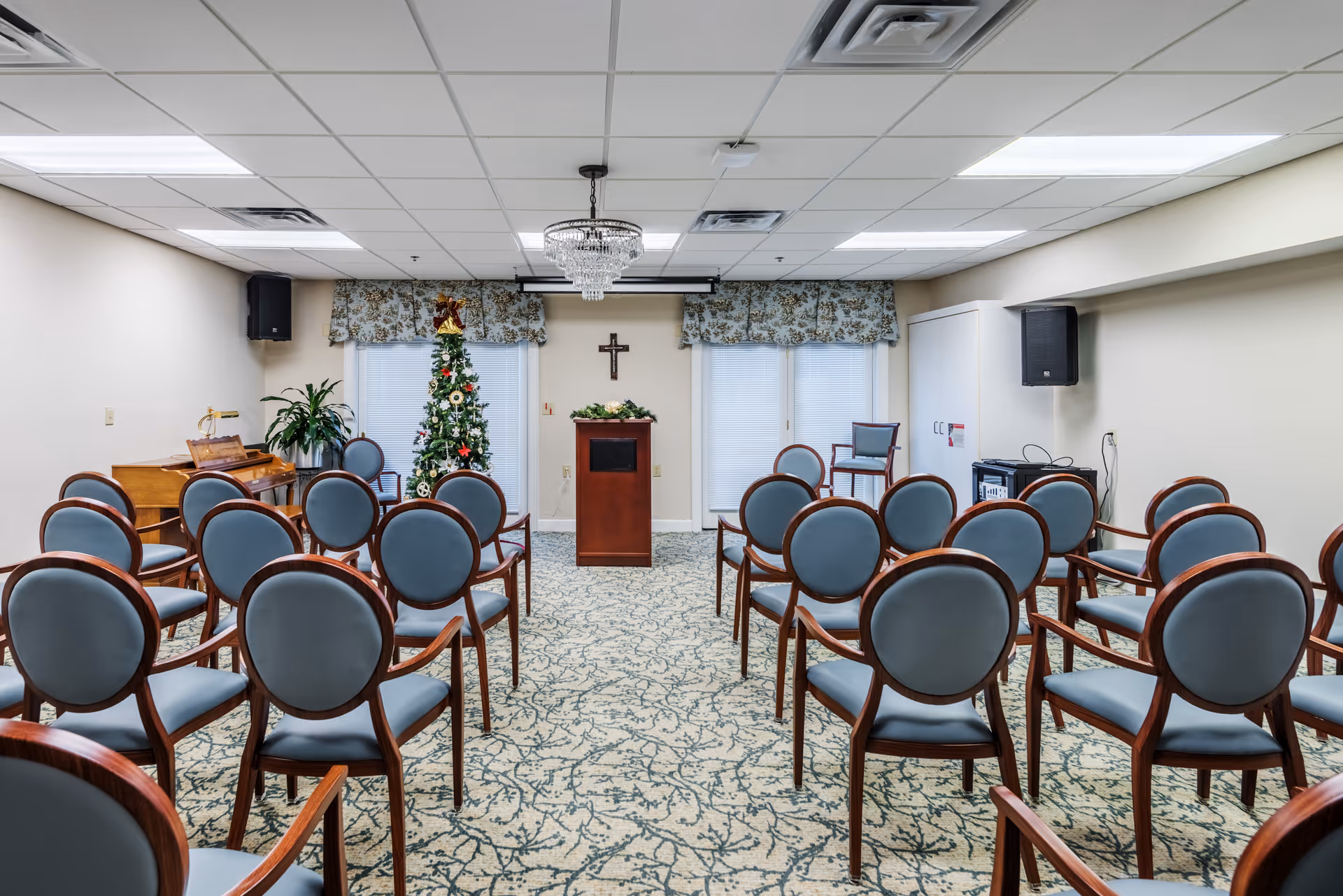 A small meeting or chapel room with rows of wooden chairs facing a lectern, a decorated Christmas tree, piano and wall cross.