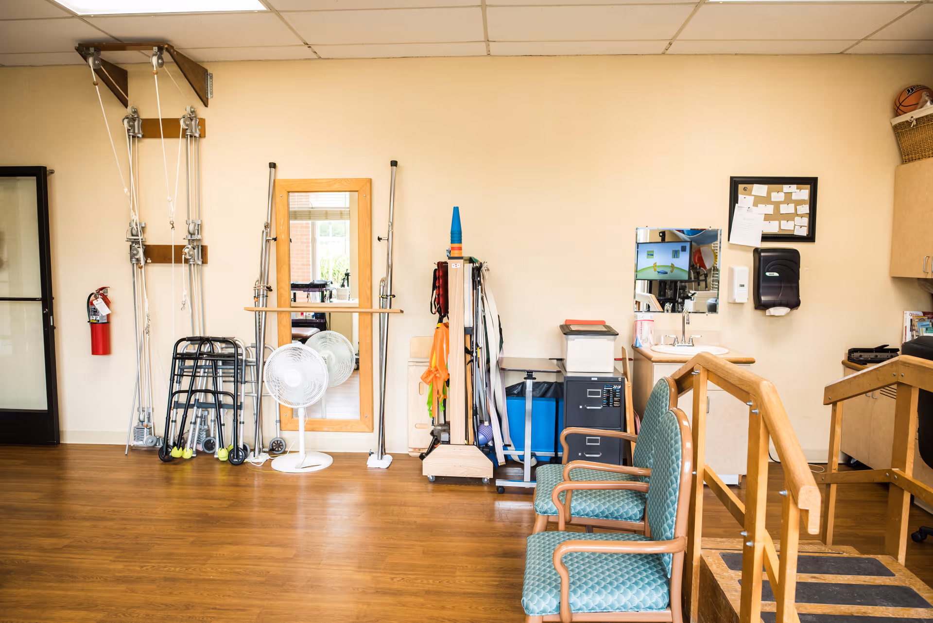 A rehabilitation therapy room with exercise equipment including pulleys, walkers, a standing mirror, a fan, and various therapy tools. There are two green cushioned chairs with wooden armrests, a small sink area with a mirror, and a wooden ramp with handrails. The room has a wooden floor and beige walls.