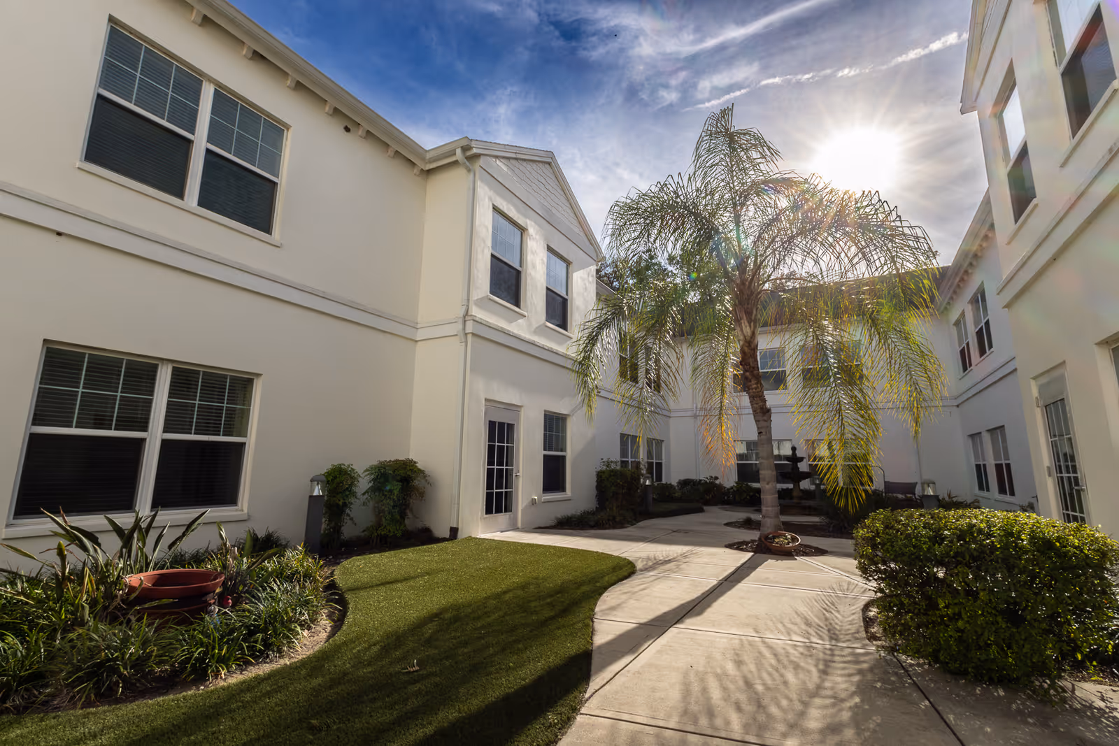 Outdoor courtyard area at The Harrison of Wildwood featuring a paved walkway, green grass, landscaped plants, a palm tree, and white two-story buildings under a partly cloudy sky with the sun shining brightly.