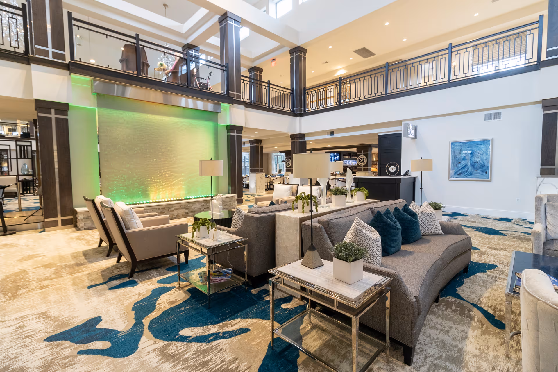 Spacious two-story senior living lounge with sofas, armchairs, side tables, a green-lit decorative water wall and patterned carpet.