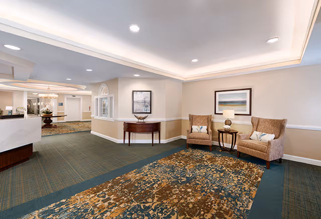 A spacious senior living facility interior with beige walls and a patterned carpet. Two upholstered armchairs with decorative pillows flank a small round table with a lamp. A framed landscape painting hangs above the chairs. In the background, there is a hallway with a reception desk, a wooden console table, and additional seating areas.