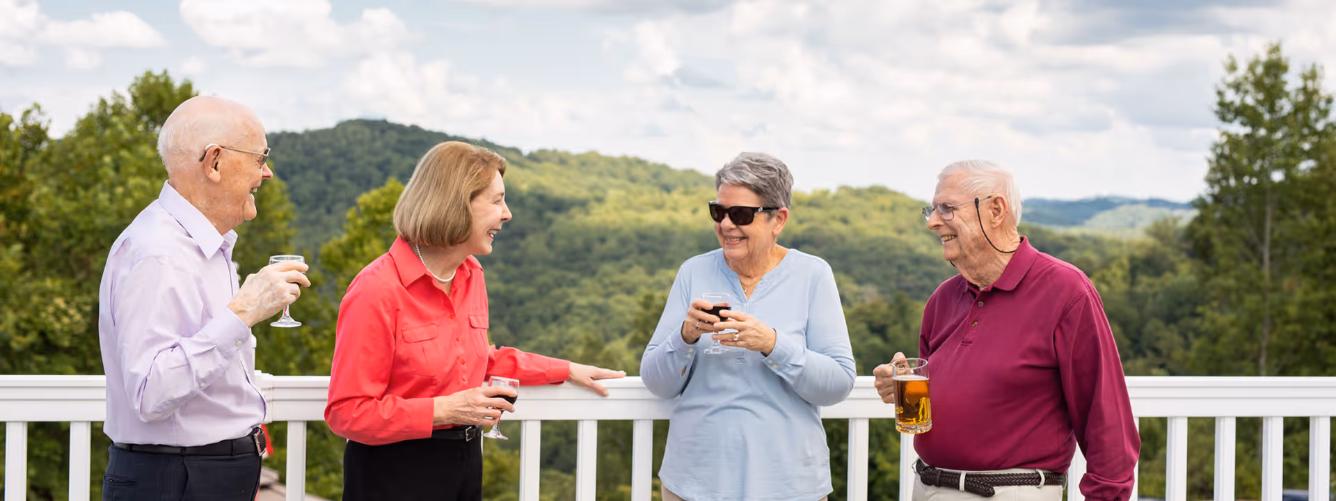 Four elderly people standing outdoors on a balcony with a white railing, holding drinks and smiling while engaging in conversation. Behind them is a scenic view of green forested hills under a partly cloudy sky.