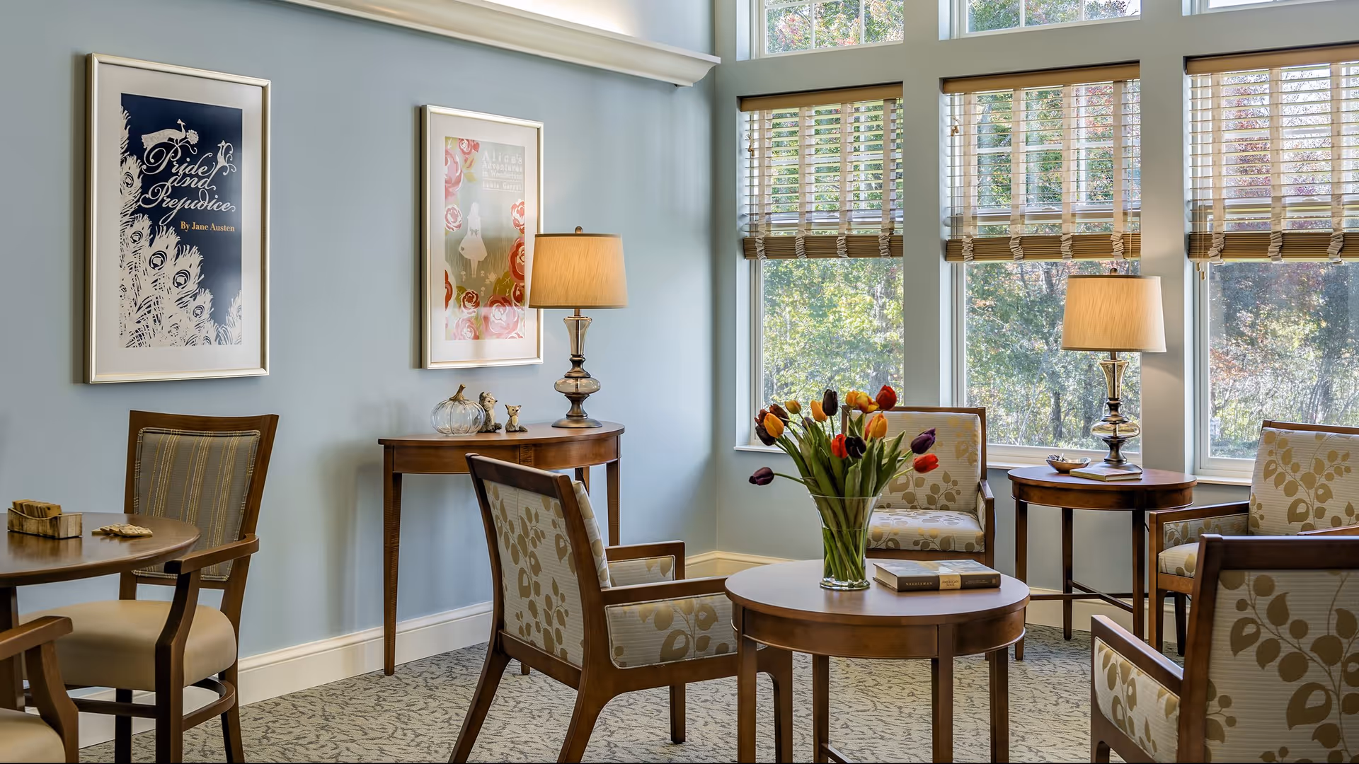 A cozy sitting area with wooden chairs upholstered in beige fabric with leaf patterns, a round wooden table with a vase of colorful tulips, two side tables with lamps, and large windows with wooden blinds letting in natural light. The walls are painted light blue and decorated with framed artwork.