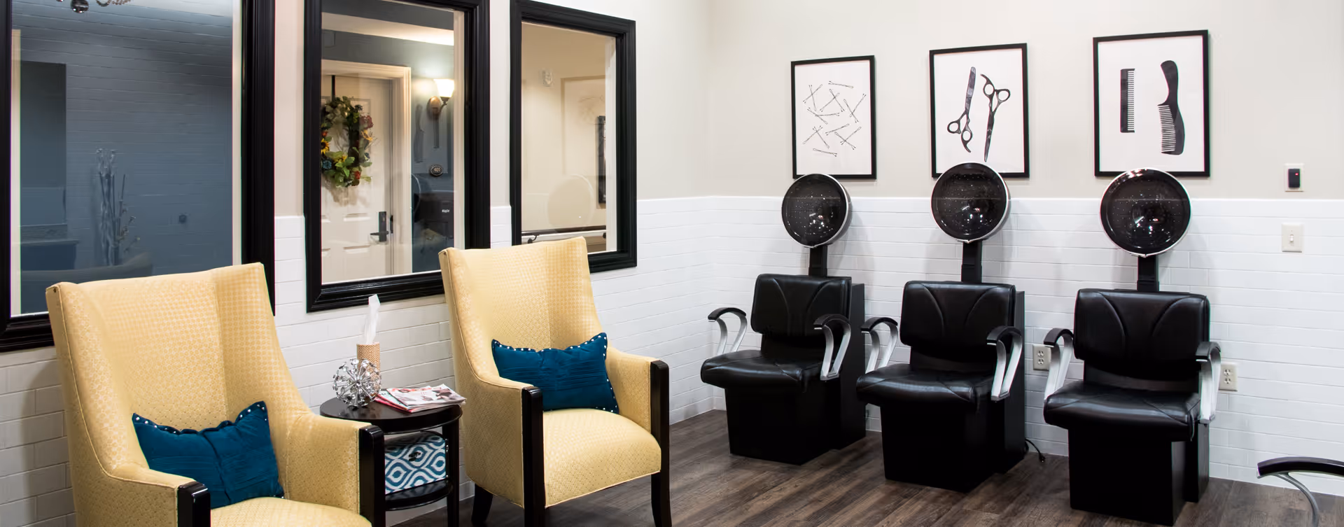 Interior of a salon area with three black hair drying chairs lined up against a wall. Above the chairs are three framed pictures depicting hair styling tools such as scissors, combs, and hairpins. To the left, there are two yellow armchairs with blue cushions and a small round table between them holding magazines and a tissue box. The walls are white with a tiled lower half and the floor is wooden.