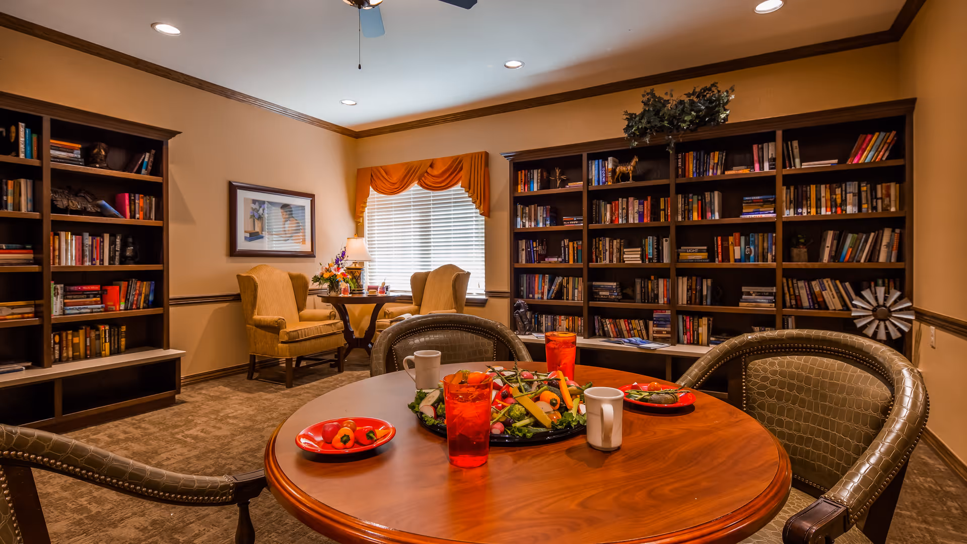 A cozy common room with a round wooden table set with plates of food and drinks. Surrounding the table are cushioned chairs with a textured pattern. In the background, there are large bookshelves filled with books, two upholstered armchairs near a window with orange valance curtains, and a framed picture on the wall. The room has warm lighting and a ceiling fan.
