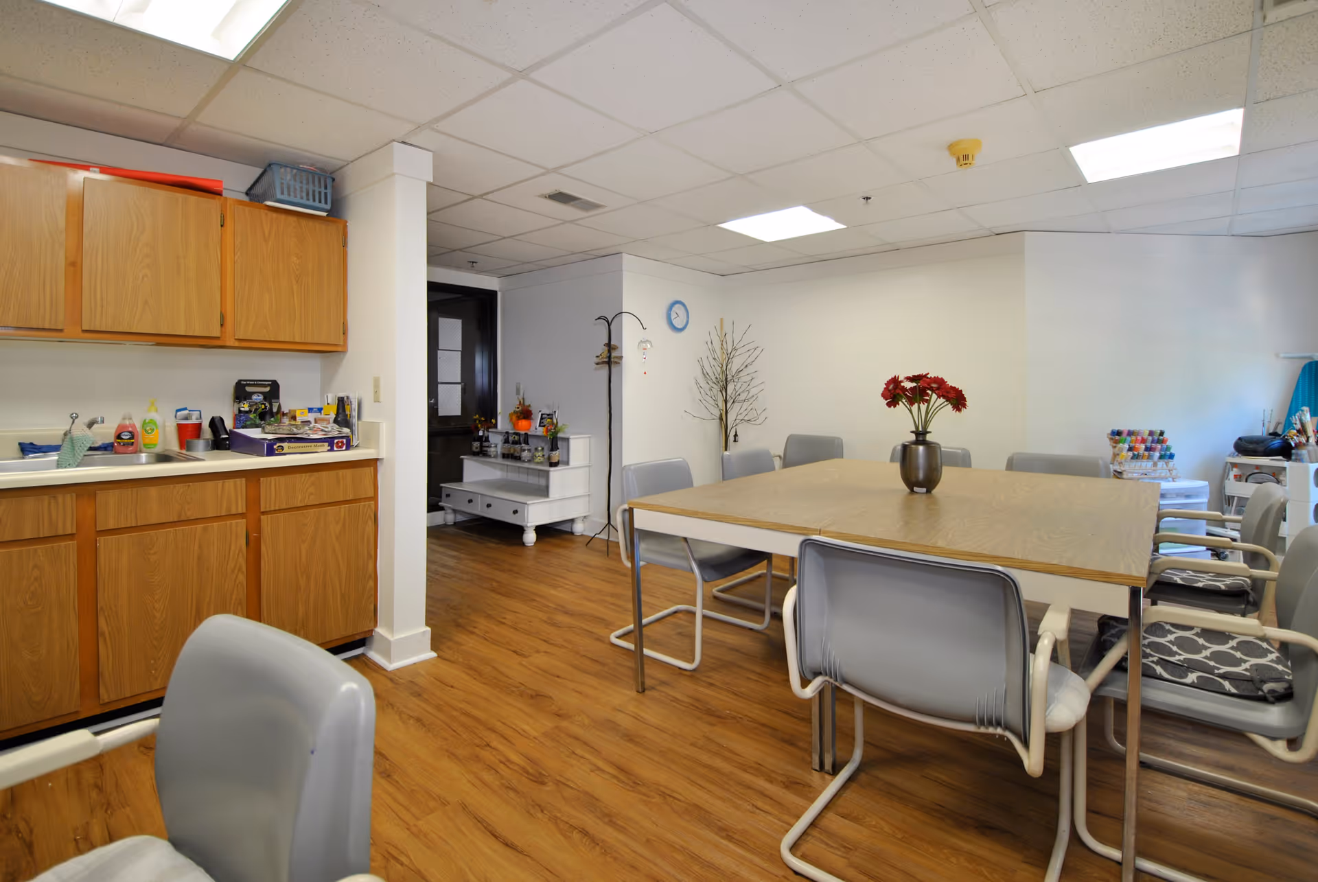 A room with a wooden floor and a ceiling with fluorescent lights. There is a rectangular table in the center surrounded by gray chairs, some with cushions. On the table is a vase with red flowers. To the left, there is a kitchenette area with wooden cabinets, a sink, and various items on the counter. In the background, there is a small white cabinet with decorations and a coat rack. The walls are white and there is a clock on one wall.
