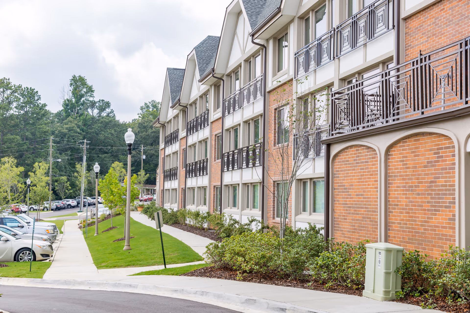 Exterior view of a three-story brick senior living building with balconies, landscaped sidewalk, lampposts, and parked cars.