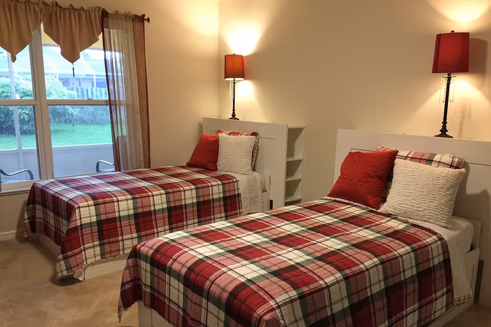A bedroom with two single beds, each covered with a red, white, and green plaid blanket. Each bed has a red pillow and a white textured pillow. There are two bedside lamps with red lampshades mounted on the wall above the headboards. A window with brown curtains is visible on the left side, showing greenery outside.