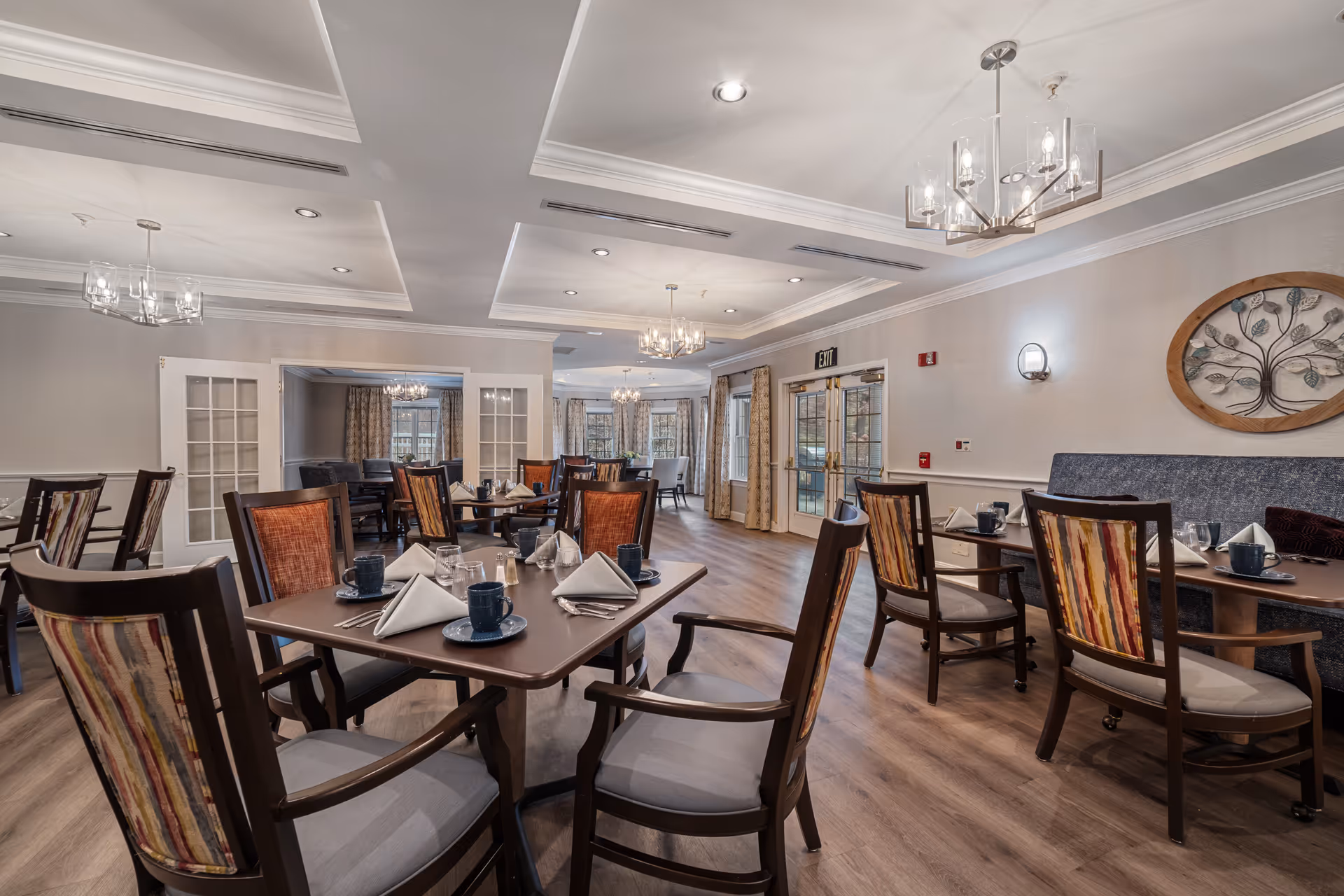 A well-lit dining room in a senior living facility with multiple tables set with blue cups, plates, silverware, and folded white napkins. The room features wooden chairs with colorful striped upholstery, hardwood floors, chandeliers, and large windows with curtains allowing natural light to enter. There is a decorative wall art piece and an exit door visible in the background.