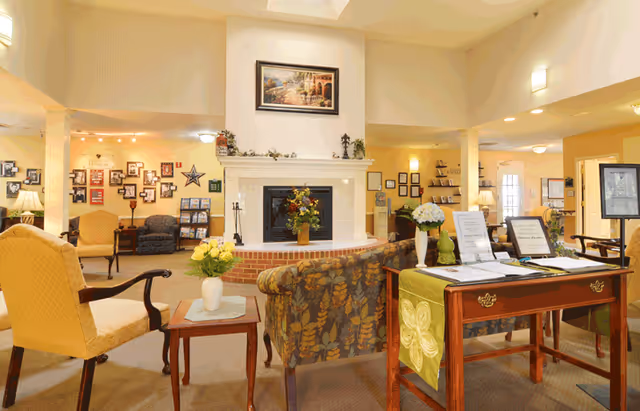 A spacious and warmly lit senior living facility common area featuring comfortable upholstered chairs and sofas arranged around a central fireplace with a floral arrangement on the mantel. The room has high ceilings with wall sconces, framed artwork above the fireplace, and various informational displays and decorations on the walls. A wooden table with informational brochures and a green table runner is positioned in the foreground.