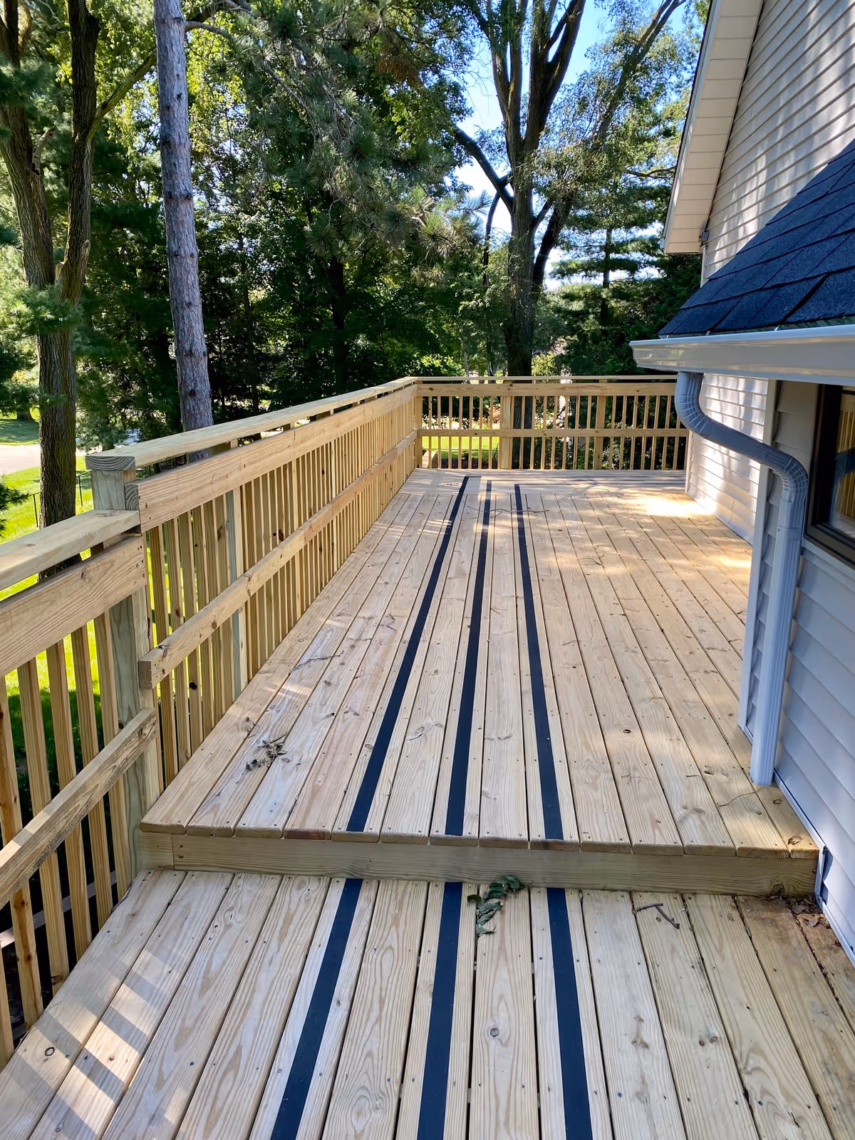 A wooden outdoor deck attached to a building with light-colored siding. The deck has black anti-slip strips on the floor and wooden railings. Trees and greenery are visible in the background under a clear blue sky.