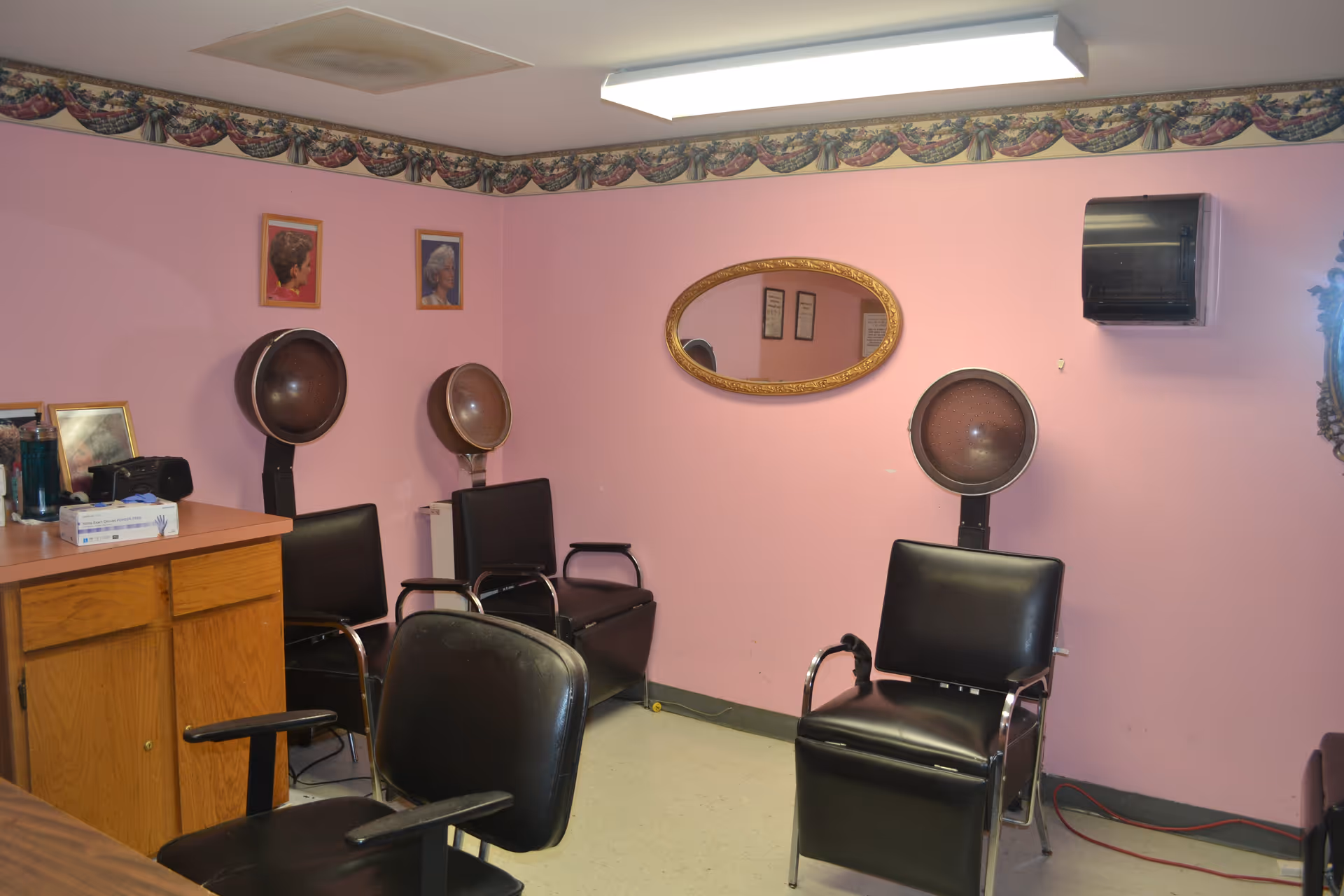 Interior of a small hair salon or beauty room with pink walls, three black salon chairs with attached hair dryers, a wooden cabinet with various items on top, an oval mirror on the wall, and two framed pictures of hairstyles.