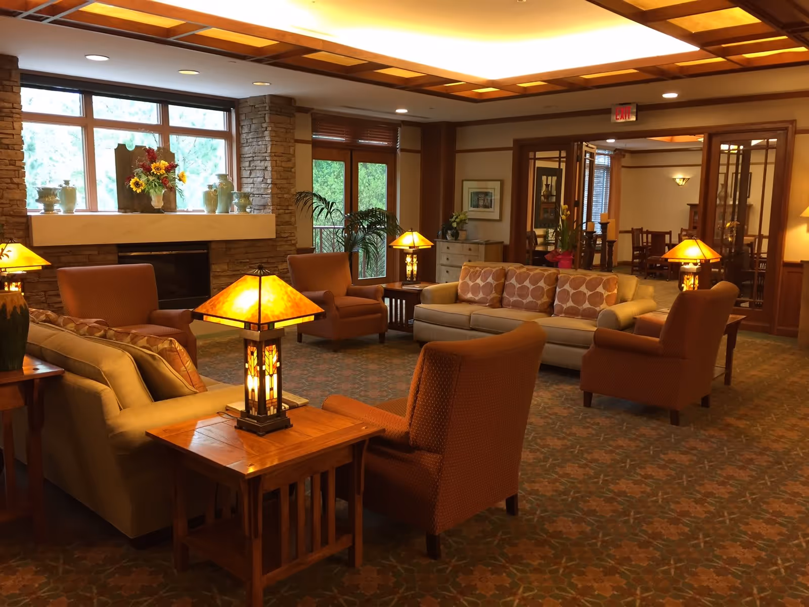 A cozy living room area with beige and orange upholstered chairs and sofas arranged around wooden side tables with warm glowing lamps. The room features a stone fireplace with decorative vases and flowers on the mantel, large windows letting in natural light, and a patterned carpet. In the background, there is a dining area visible through glass doors.