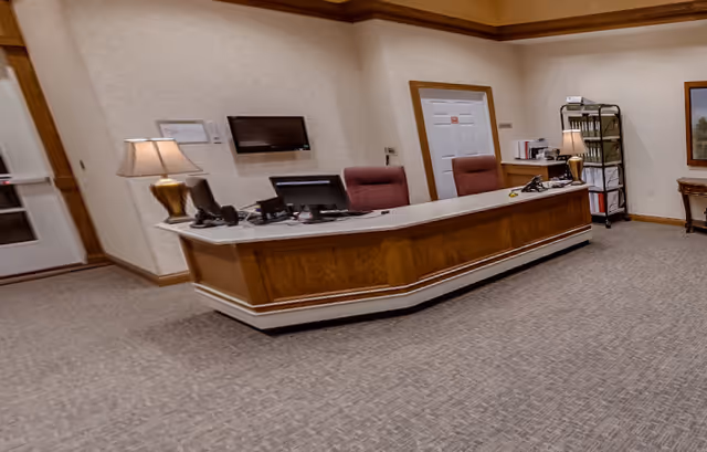 Reception desk in a carpeted lobby with chairs, desk lamps, computer monitors and a shelving cart.