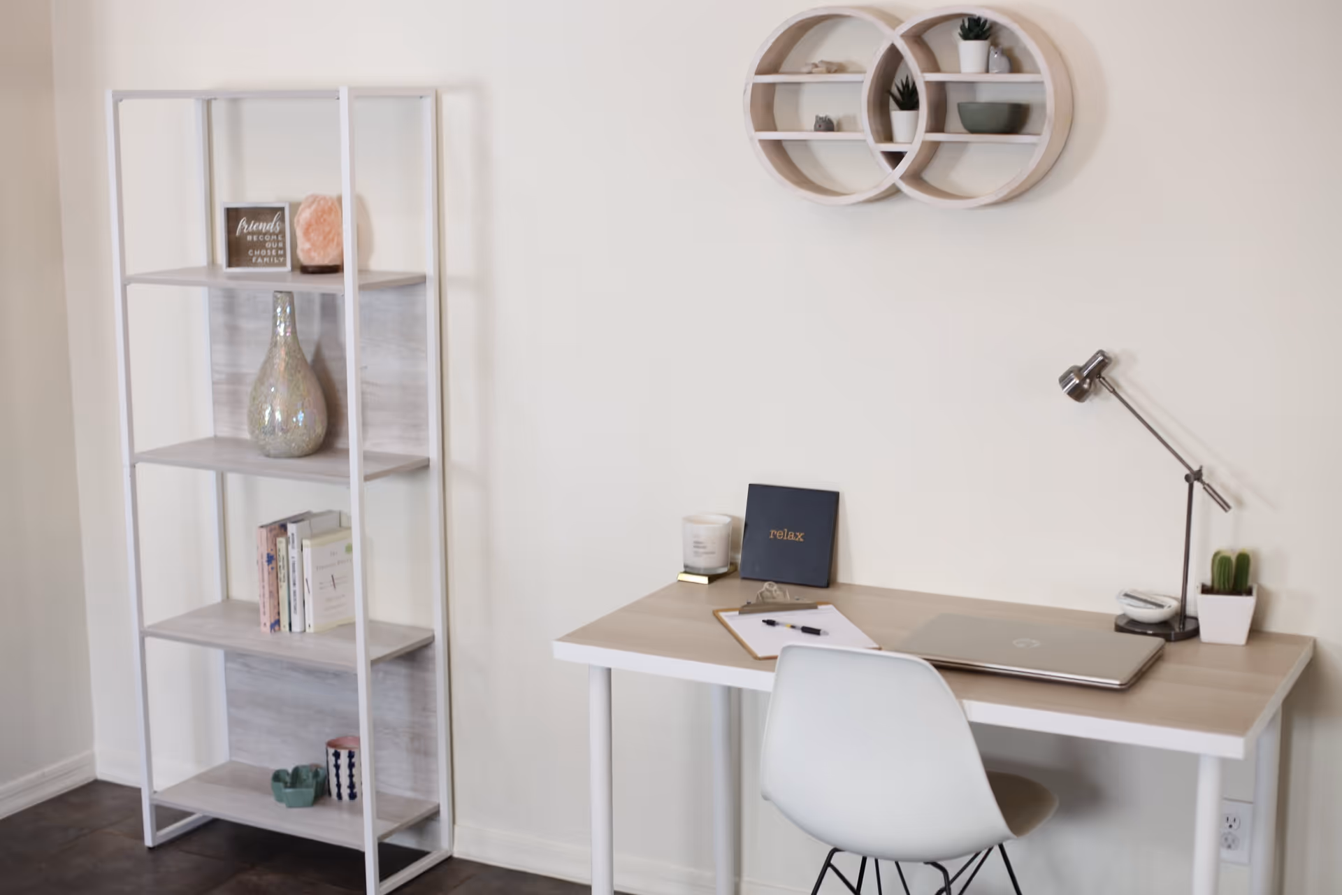 A minimalist workspace featuring a light wood desk with a closed laptop, a clipboard with a pen, a candle, a small sign that says 'relax', a modern desk lamp, and a small potted cactus. To the left is a white and light wood open shelving unit with decorative items including books, a vase, and small plants. Above the desk is a circular wall shelf with small plants and decor items.