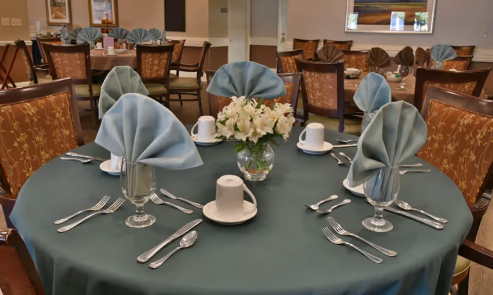 Dining room table with a green tablecloth, folded napkins in goblets, place settings and a small vase of white flowers.