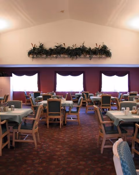 Dining room with multiple tables covered with light blue tablecloths and wooden chairs with green cushions. The room has large windows with white curtains and maroon valances. Above the windows, there is a shelf with green plants. The carpet is patterned in shades of red and brown.