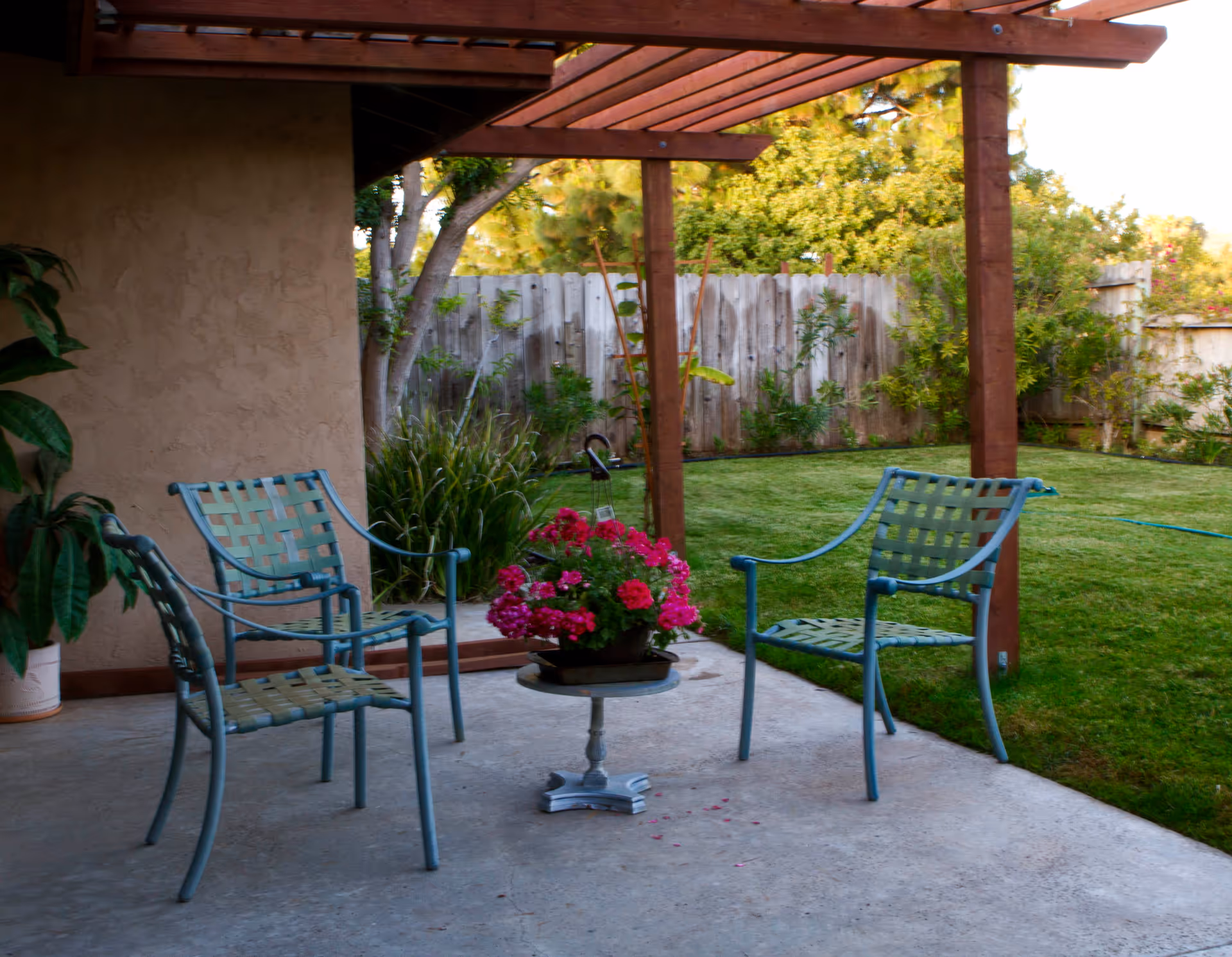 Outdoor patio area with three metal chairs arranged around a small round table holding a pot of pink flowers. The patio is covered by a wooden pergola and overlooks a grassy backyard with trees and a wooden fence.