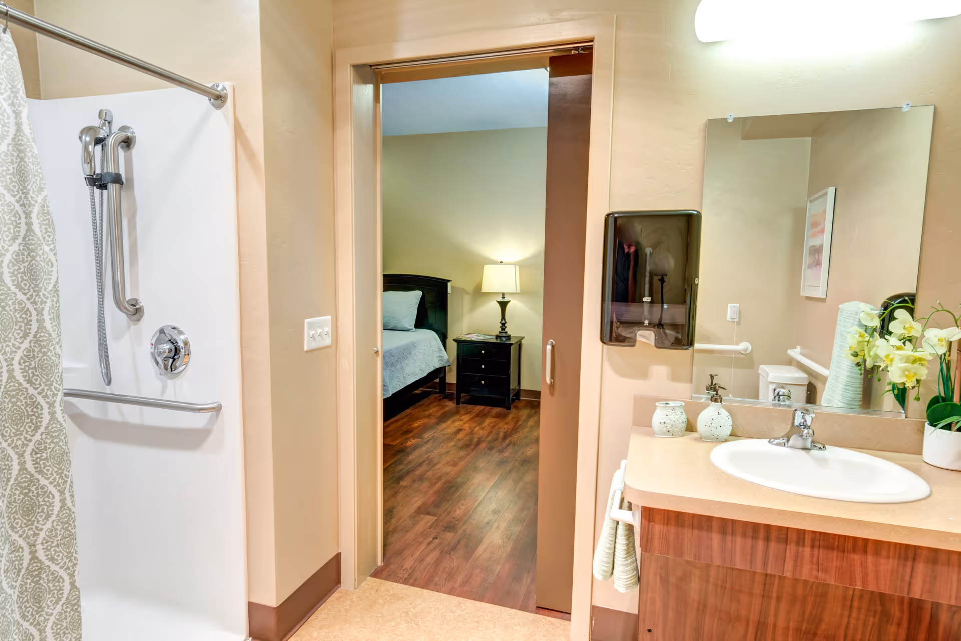 View of a bathroom with a shower area on the left, a sink with a countertop and mirror on the right, and an open door leading to a bedroom with a bed, nightstand, and lamp visible.