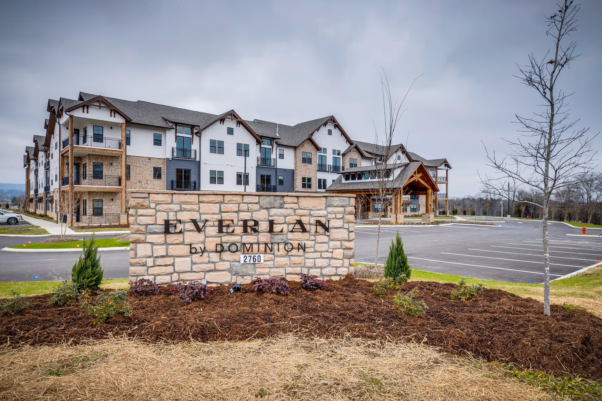 Exterior view of Everlan by Dominion senior living facility in Hixson, showing a large stone sign with the facility name and a multi-story building with balconies and a covered entrance in the background under a cloudy sky.