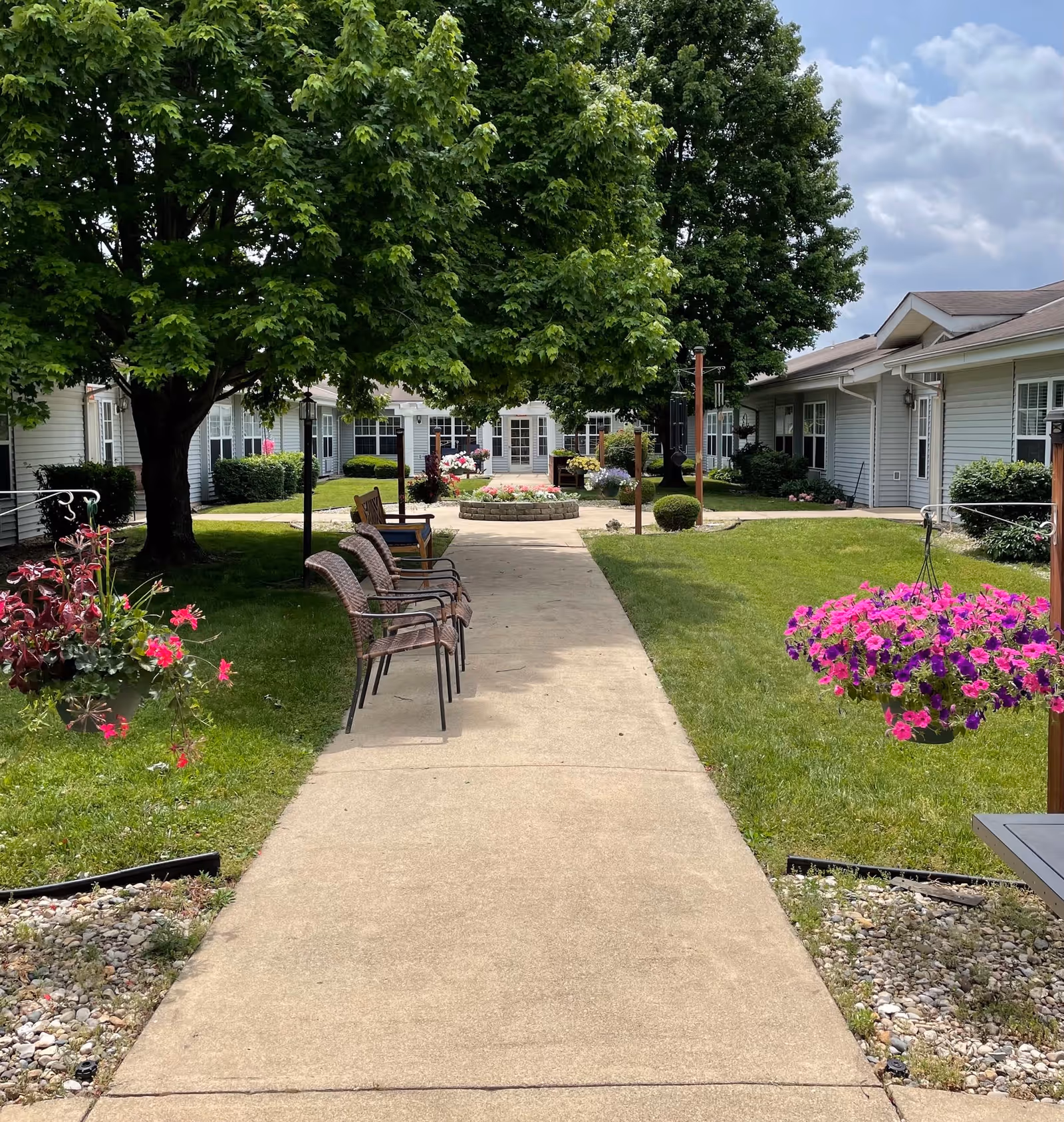 A peaceful outdoor courtyard at Cedar Creek of Logansport featuring a concrete walkway lined with chairs on one side, lush green grass, hanging flower baskets with pink and purple flowers, large leafy trees providing shade, and single-story residential buildings with white siding surrounding the area under a partly cloudy sky.