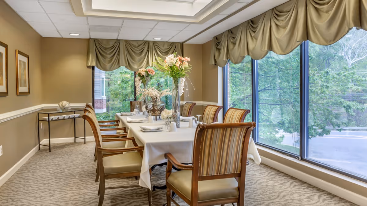A formal dining room with a long table covered with a white tablecloth, set with plates, glasses, and floral centerpieces. The room has large windows with green drapes, allowing natural light to fill the space. There are several wooden chairs with striped upholstery around the table, and framed artwork on the beige walls.