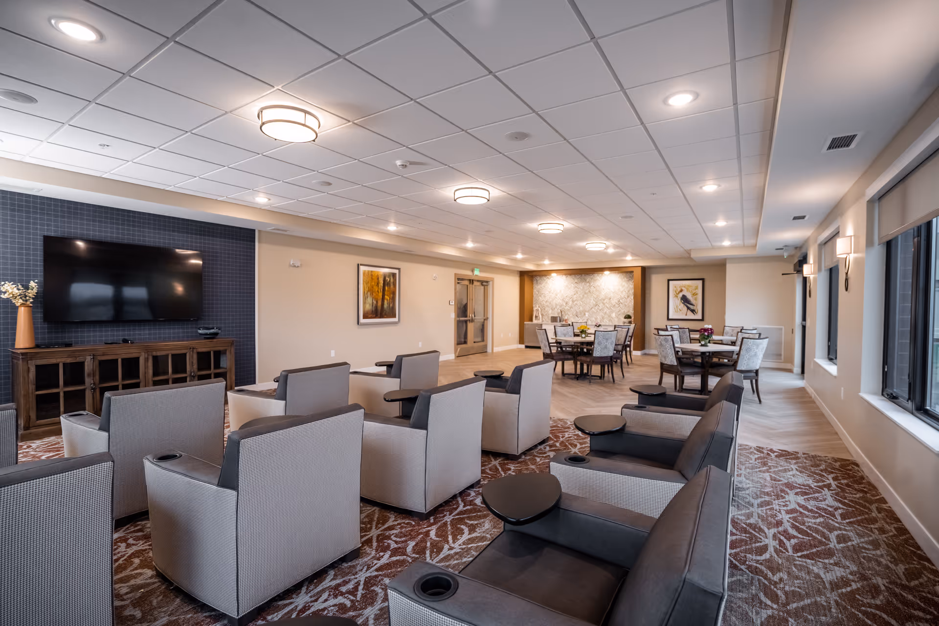 A spacious and well-lit common area with multiple comfortable armchairs arranged in rows facing a large flat-screen TV mounted on a dark accent wall. The room features a patterned carpet, several round tables with chairs in the background, framed artwork on the walls, and large windows allowing natural light to enter.