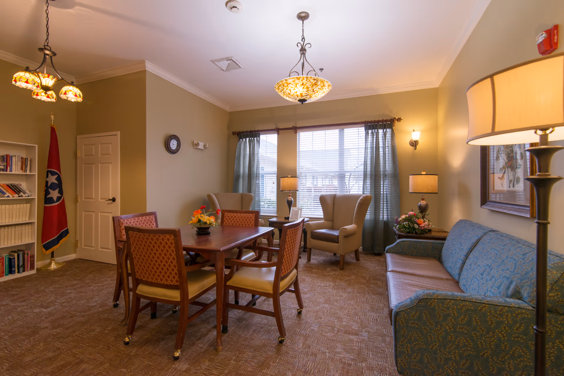 Cozy senior living common room with a central dining table and chairs, armchairs by a window, a sofa, and multiple lamps and light fixtures.