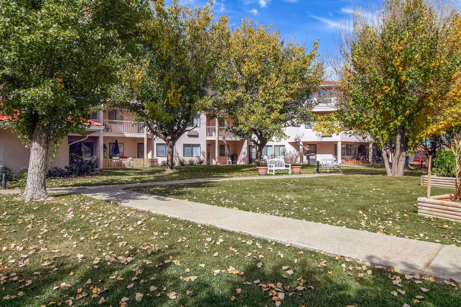 Outdoor view of Asher Point Independent Living of Amarillo showing a grassy area with a concrete walkway, several trees with green and yellow leaves, white benches, and a two-story building with balconies in the background under a blue sky with some clouds.