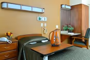 A modest senior-care bedroom with a single bed, bedside table and phone, an overbed table holding a decorative pitcher, a chair and a built-in desk with a plant.