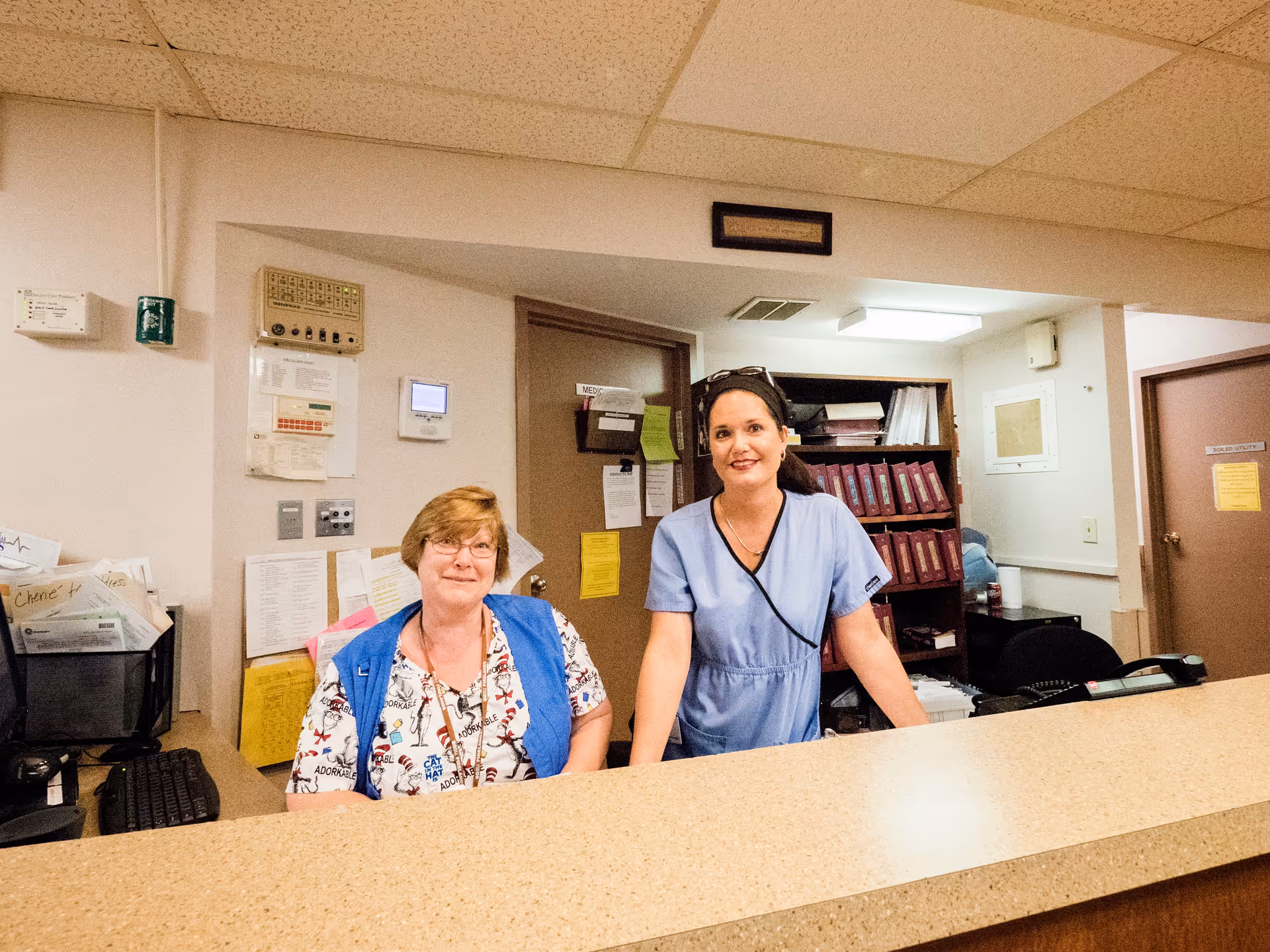 Two female healthcare workers behind a reception desk in a healthcare facility. One is seated wearing a patterned scrub top and blue vest, and the other is standing wearing light blue scrubs. Behind them are shelves with binders and office supplies, and a door labeled 'MEDIC'.