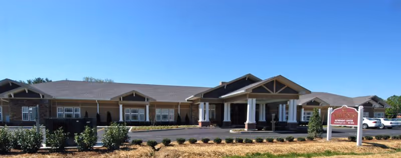 Front exterior view of Webb House Retirement Center, a single-story building with a covered entrance, multiple windows, and a landscaped area with bushes and mulch in front. Several cars are parked to the right side of the building under a clear blue sky.