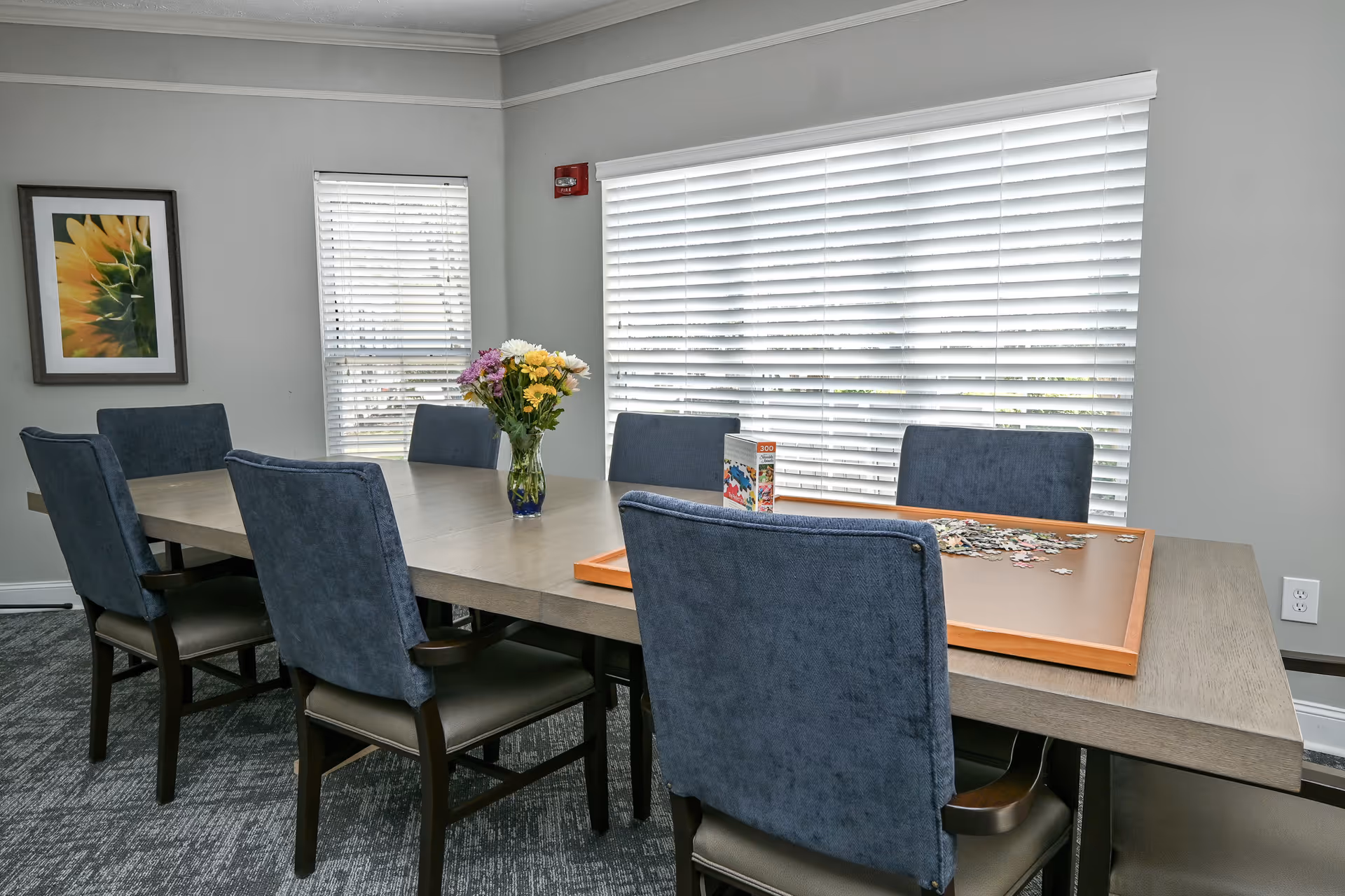 Long table with blue upholstered chairs, a vase of flowers and a partially completed puzzle near a large window with blinds.