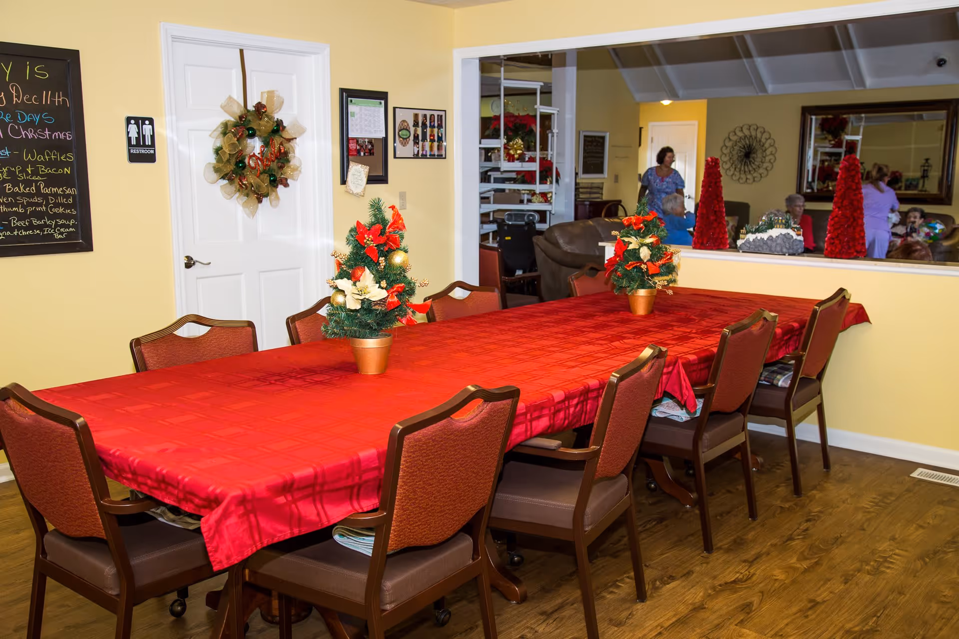 A dining room with a long table covered in a red tablecloth and decorated with small Christmas-themed centerpieces. There are eight chairs around the table. A white door with a restroom sign and a holiday wreath is on the left wall. A chalkboard menu is also visible on the left. In the background, a large mirror reflects several people sitting and standing in an adjacent room with yellow walls and wooden flooring.