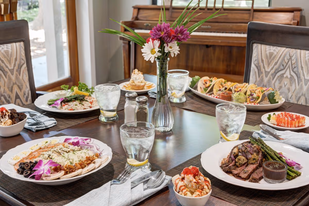 A dining table set with various plated meals including steak with asparagus, tacos, a salad, and desserts. There are glasses of water with lemon slices, silverware wrapped in napkins, a vase with purple and white flowers, and salt and pepper shakers on the table. A piano and large windows are visible in the background.