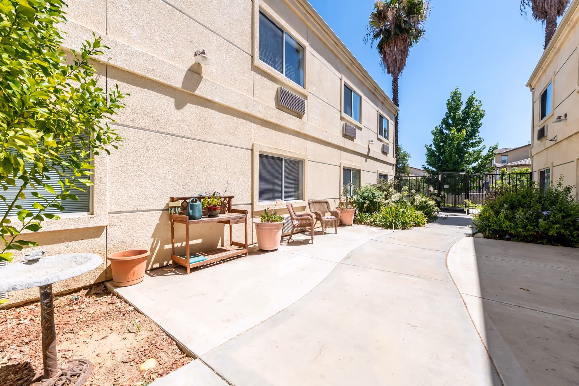 Sunlit courtyard between two beige memory-care building wings with potted plants, chairs and a gated entrance.