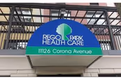 Outdoor sign for Rego Park Health Care facility mounted on a black metal fence above a concrete wall, displaying the address 11126 Corona Avenue.