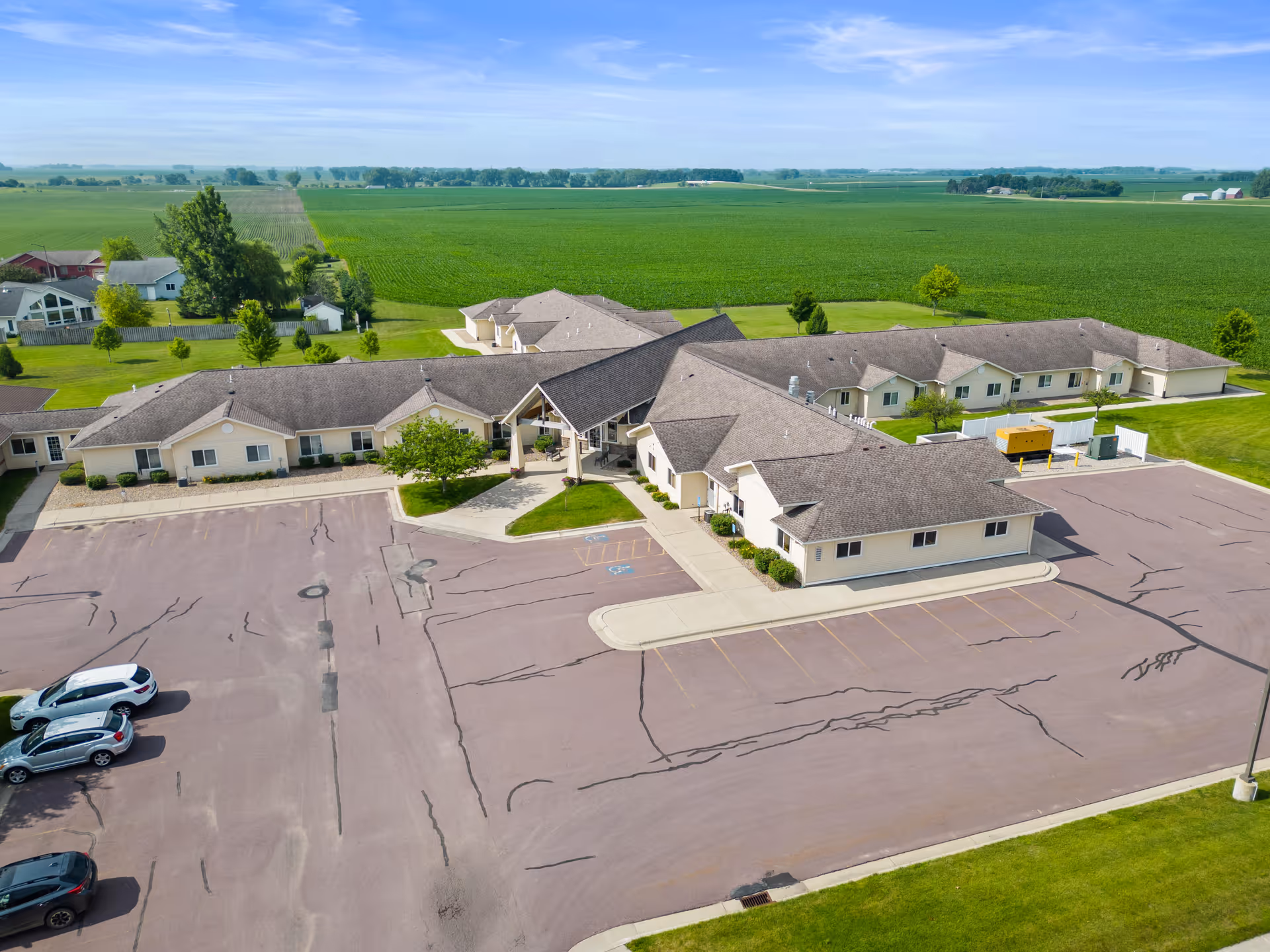 Aerial view of Good Samaritan Society - St. James - Brandt Ridge facility surrounded by green fields. The building has a beige exterior with a large parking lot in front, containing a few parked cars. The sky is clear and blue.