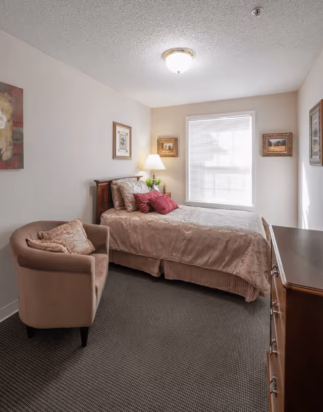 A cozy bedroom with a single bed covered in a beige bedspread and multiple pillows, including two red accent pillows. Next to the bed is a wooden nightstand with a lamp and a small plant. A beige upholstered armchair with a decorative pillow is positioned near the bed. The room has a window with blinds, framed artwork on the walls, and a wooden dresser on the right side.