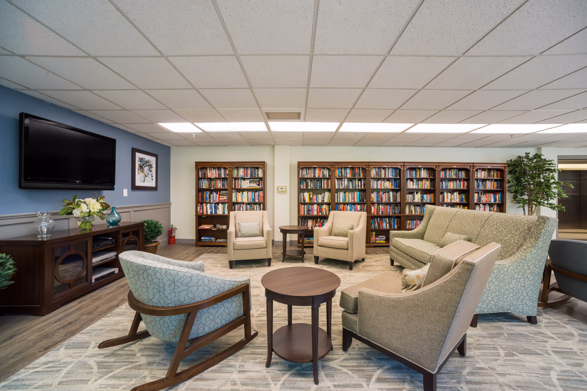 A cozy senior living room with a patterned area rug, several upholstered chairs and a sofa arranged around two round wooden tables. Behind the seating area are multiple wooden bookshelves filled with books. A flat-screen TV is mounted on a blue accent wall above a wooden media console with decorative items and plants. The ceiling has recessed lighting and a drop ceiling with white tiles.