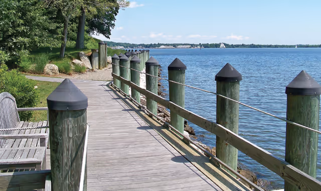 A wooden boardwalk along a waterfront with green posts topped with black caps and rope railings. There are benches on the left side and trees and grass along the shore. The water is calm under a clear blue sky with some distant land visible across the water.