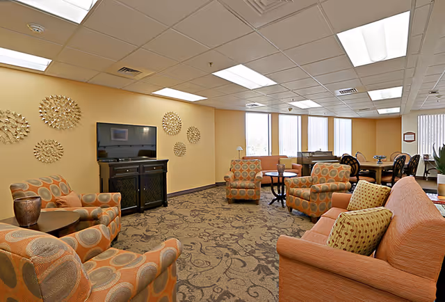 A spacious senior living facility common area with patterned armchairs and a sofa arranged around a TV on a stand. The room has beige walls decorated with circular metal wall art, large windows with vertical blinds, and a carpeted floor with a floral pattern. There is a dining table with chairs in the background.