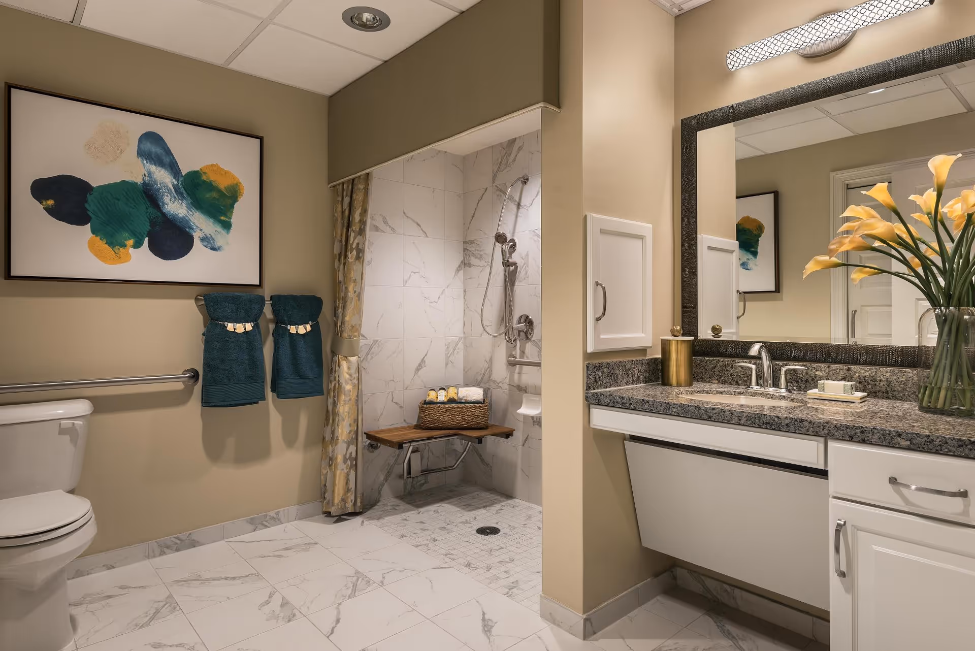 A modern bathroom featuring a toilet, a walk-in shower with a wooden bench and handheld showerhead, a granite countertop with a sink, a large mirror, and a vase of yellow calla lilies. The walls are beige, and there is abstract artwork above two teal towels hanging on a towel bar.