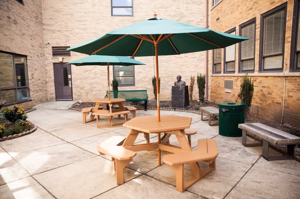 Outdoor courtyard area with two wooden picnic tables each shaded by a large green umbrella. There are two wooden benches, a green trash can, and a bust statue on a pedestal against the brick building walls surrounding the courtyard.